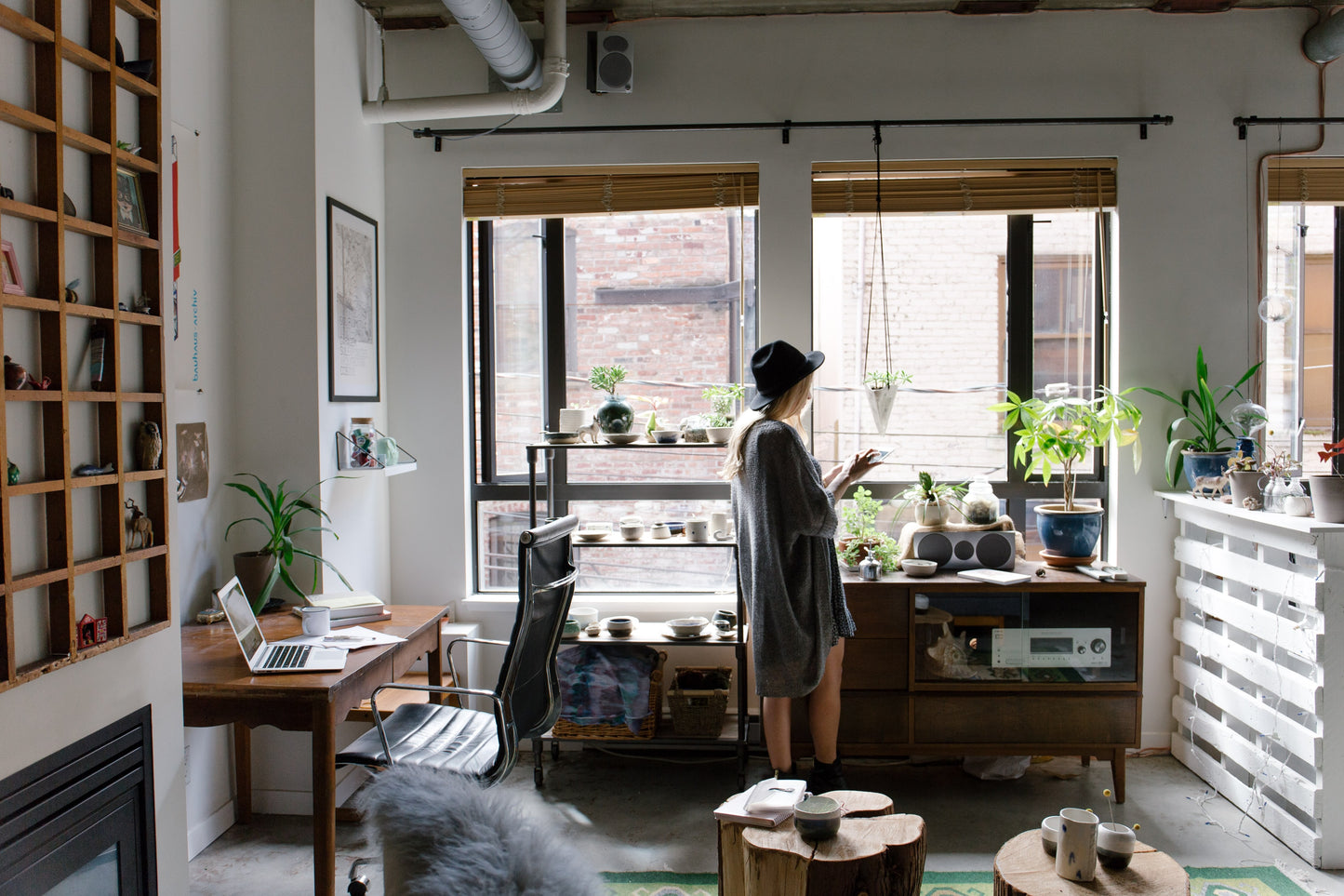 A young woman wearing a hat stands in her apartment filled with natural light and plants.