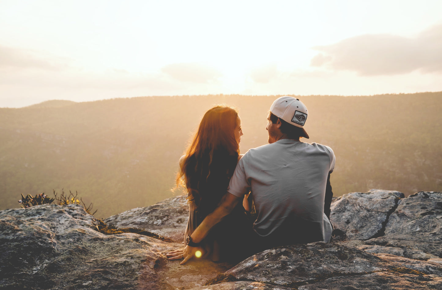 A young couple sits on a rock near a mountain range at sunset, staring into one another's eyes.