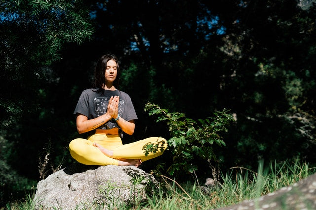 A young woman sits atop a rock outdoors with legs folded and hands in prayer position, meditating.