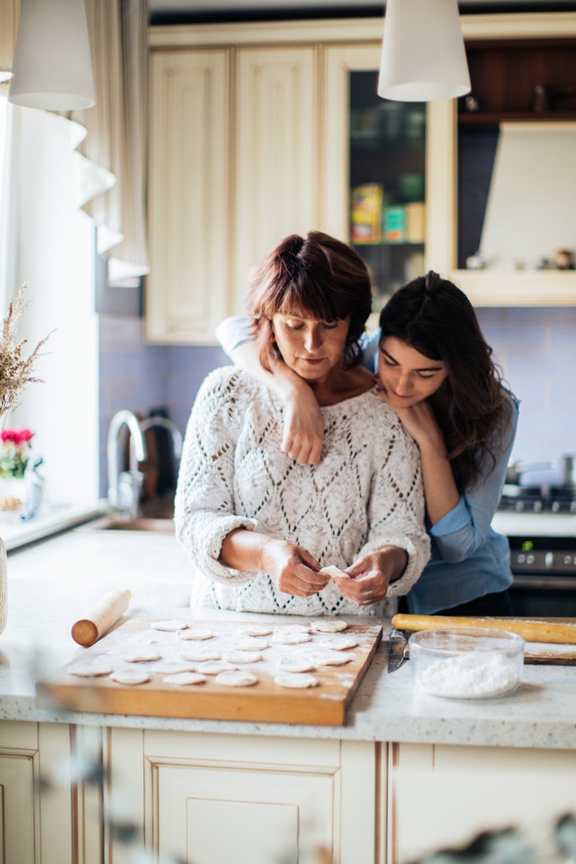 A mother bakes cookies in the kitchen as her adult daughter watches with her arms around Mom's shoulders.