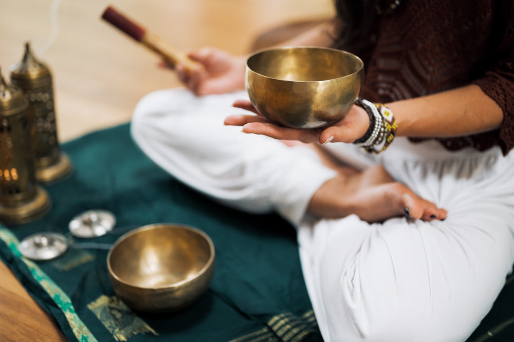A woman sits in lotus position holding a singing bowl, with candles and tingsha bells laid out nearby.