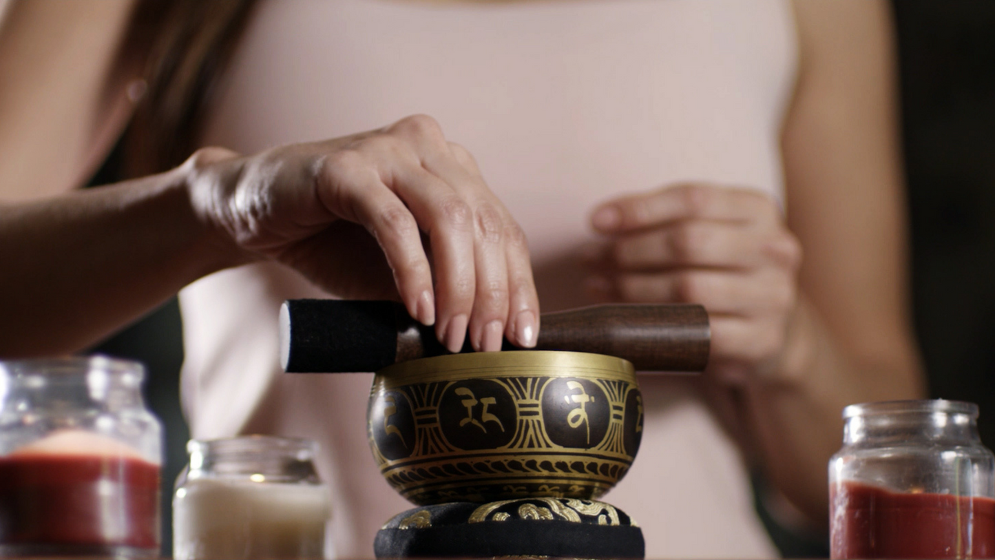 A Silent Mind singing bowl sits atop a black and gold cushion as a woman balances the striker on top, surrounded by candles.