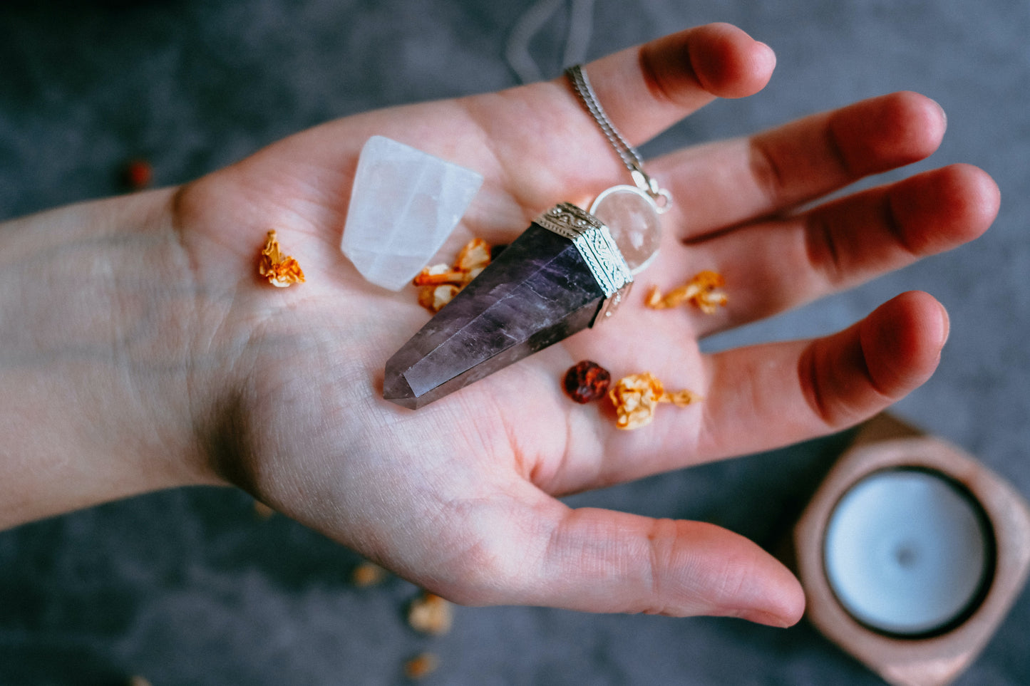 An amethyst pendulum and a small piece of quartz sit on an open palm with a tea candle in the background.