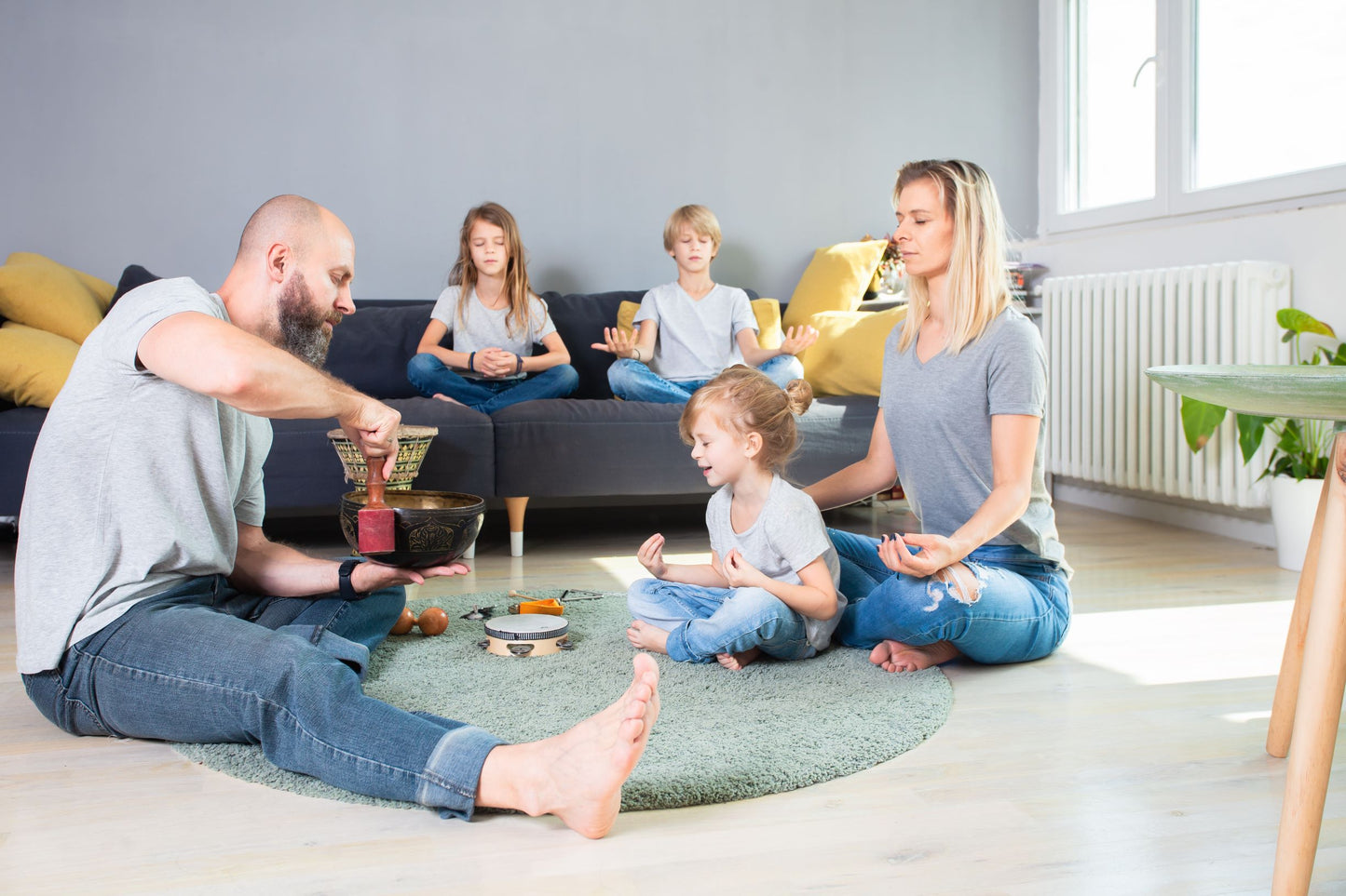 A father plays the singing bowl at home while three children and a woman practice meditation postures. 
