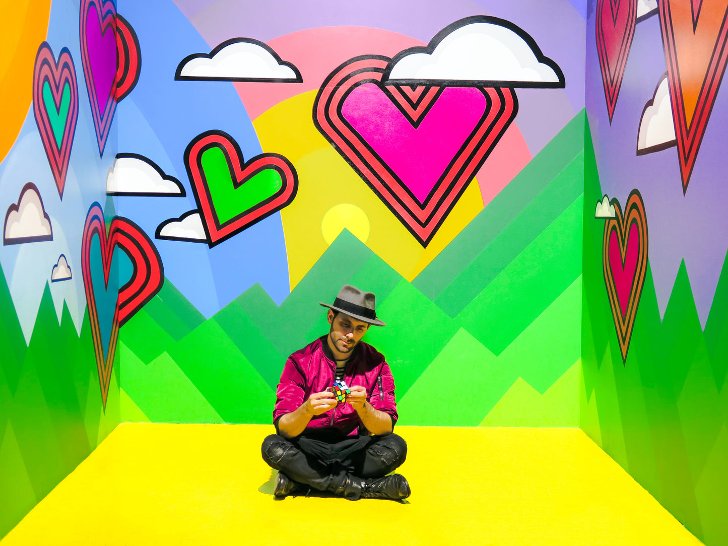 A man in a brightly colored jacket sits alone in a brightly-painted room, solving a Rubik's cube.