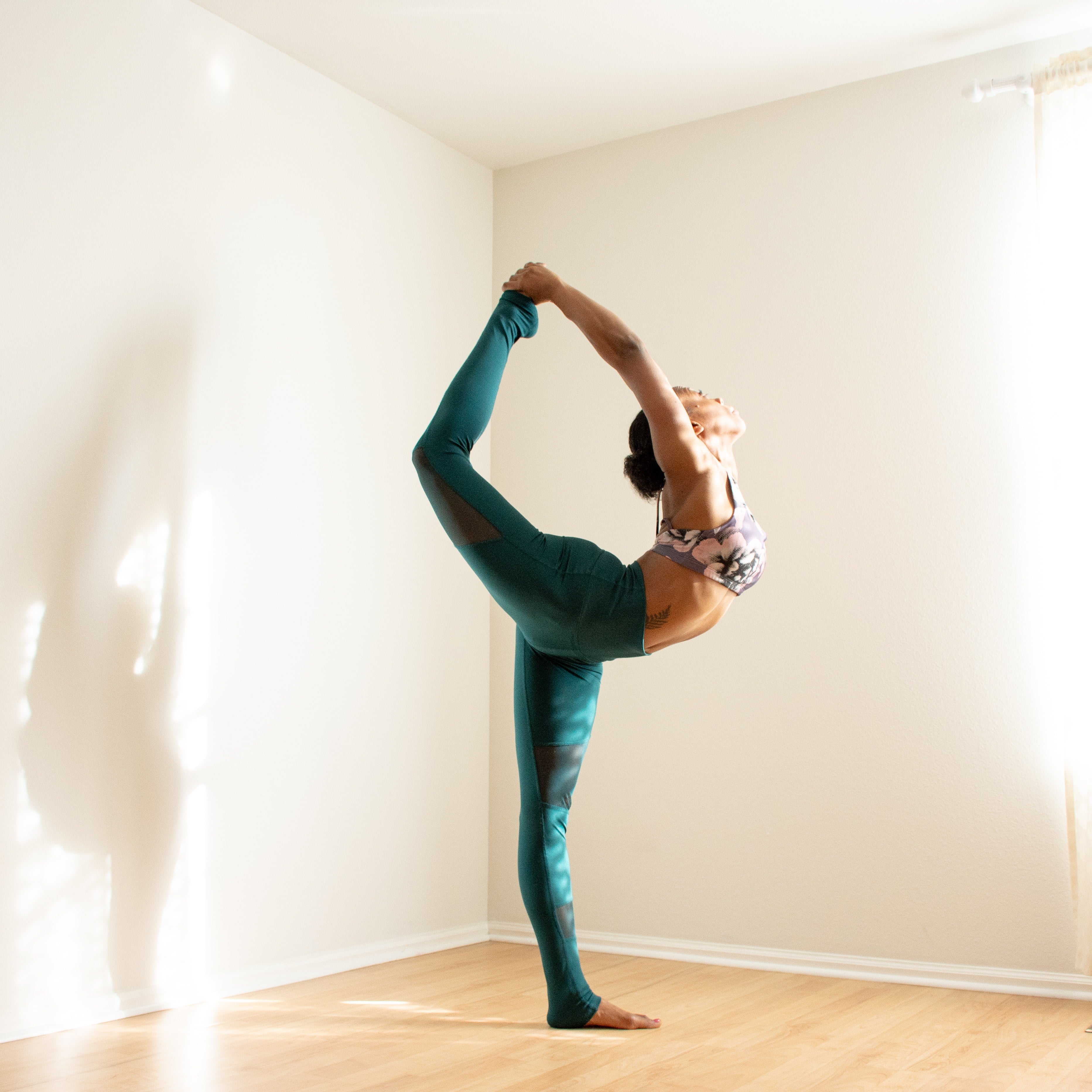 A flexible yoga practitioner performs dancer's pose in the corner of a neutral, empty room.