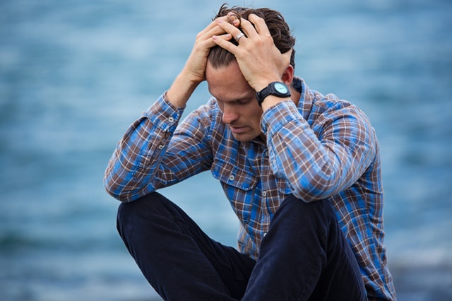 A man with health anxiety sits in front of a body of water seeming distraught with his head in his hands.