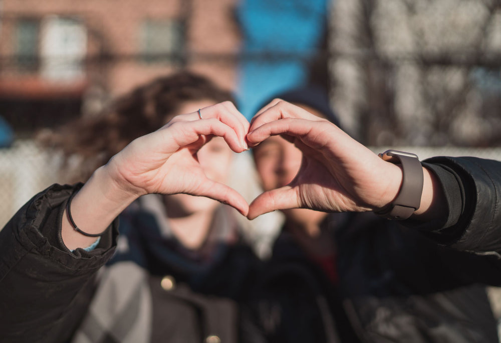 A man and woman sitting outside make a heart shape together with their hands.