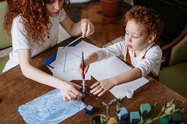 A young mother with red hair assists her child with an art project at their table.
