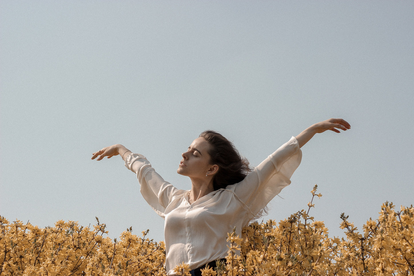 A young woman with arms outstretched and eyes closed stands in a field of flowers.