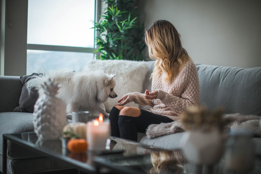 A woman sits on her couch at home playing with a fluffy white dog as a candle burns on the coffee table.