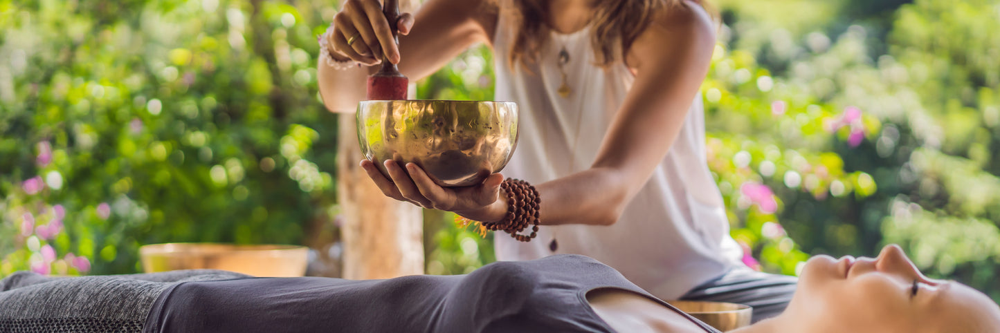 A female sound healer creates a sound bath by playing a singing bowl above a female patient lying down. 