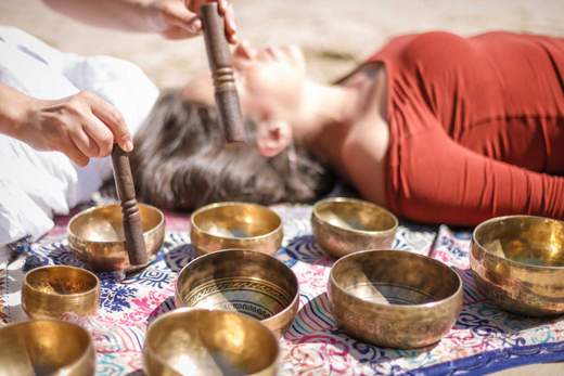 A woman lies down for sound therapy as someone plays several small singing bowls laying beside her.