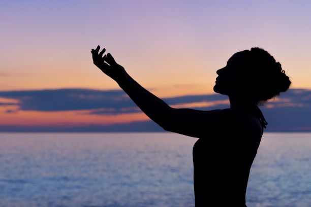 A silhouette of a woman with her arms aloft in front of an ocean at sunset.