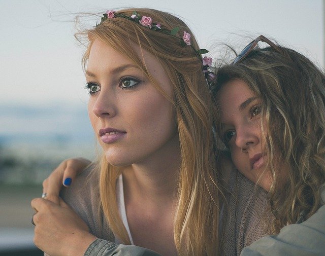 A young woman wearing a flower crown sits in the embrace of her friend.