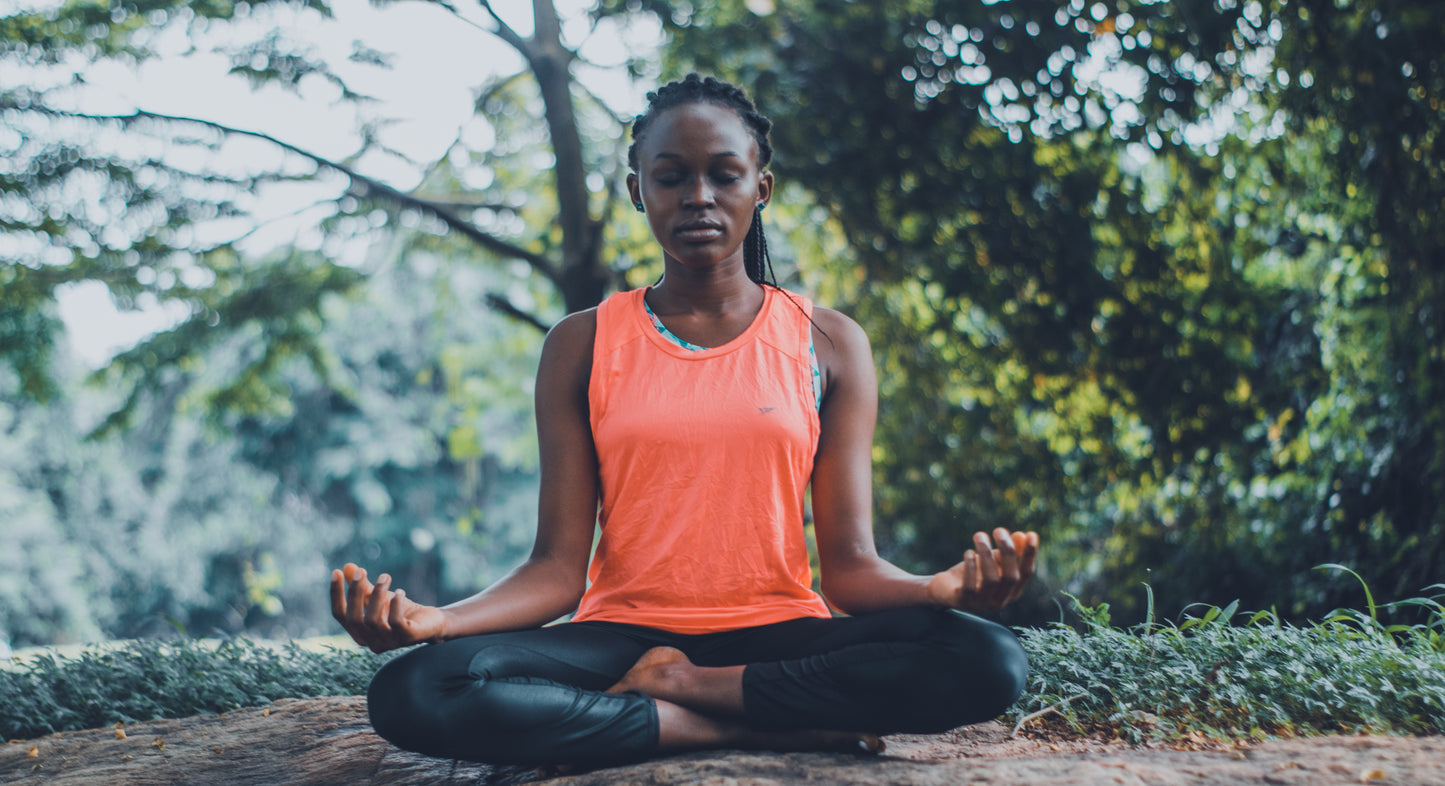 A young black woman sits outside in meditation posture with her eyes closed. 