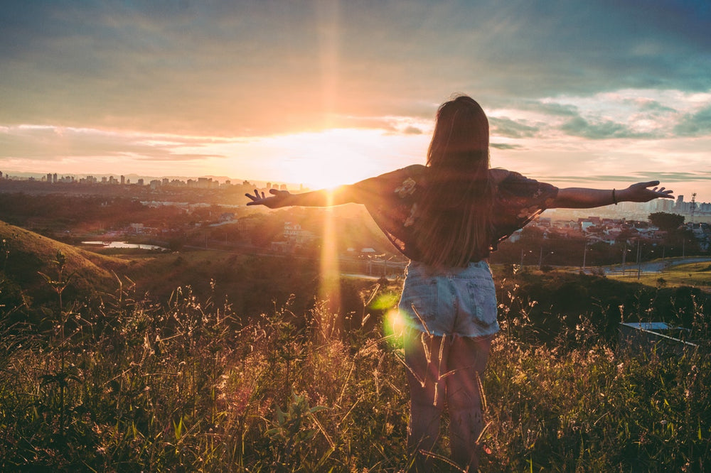 A young woman stands with her arms outstretched in a meadow at sunset, looking out at a cityscape.
