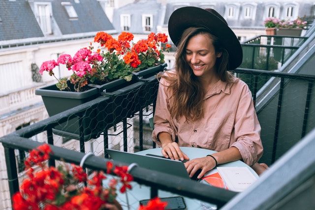 A chic woman in a wide-brimmed hat sits on a patio, smiling and working on her laptop.