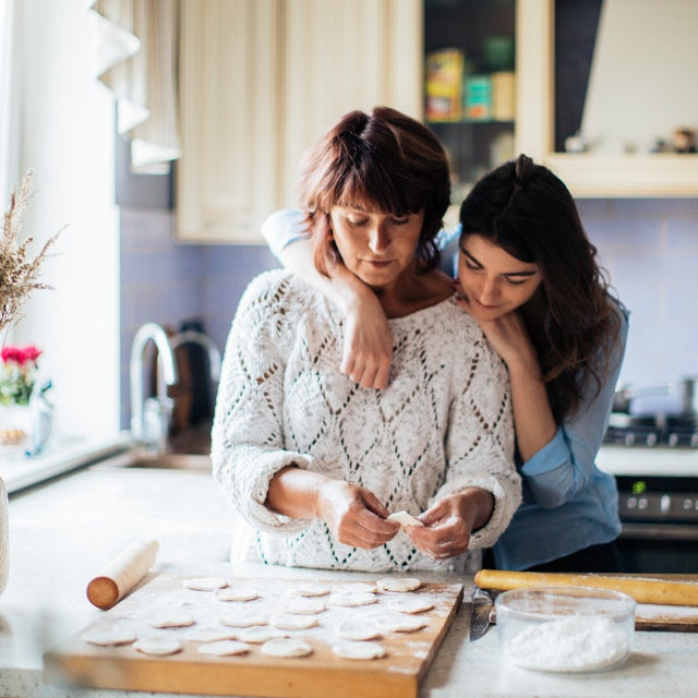 A mother bakes cookies in the kitchen as her adult daughter watches with her arms around Mom's shoulders. 
