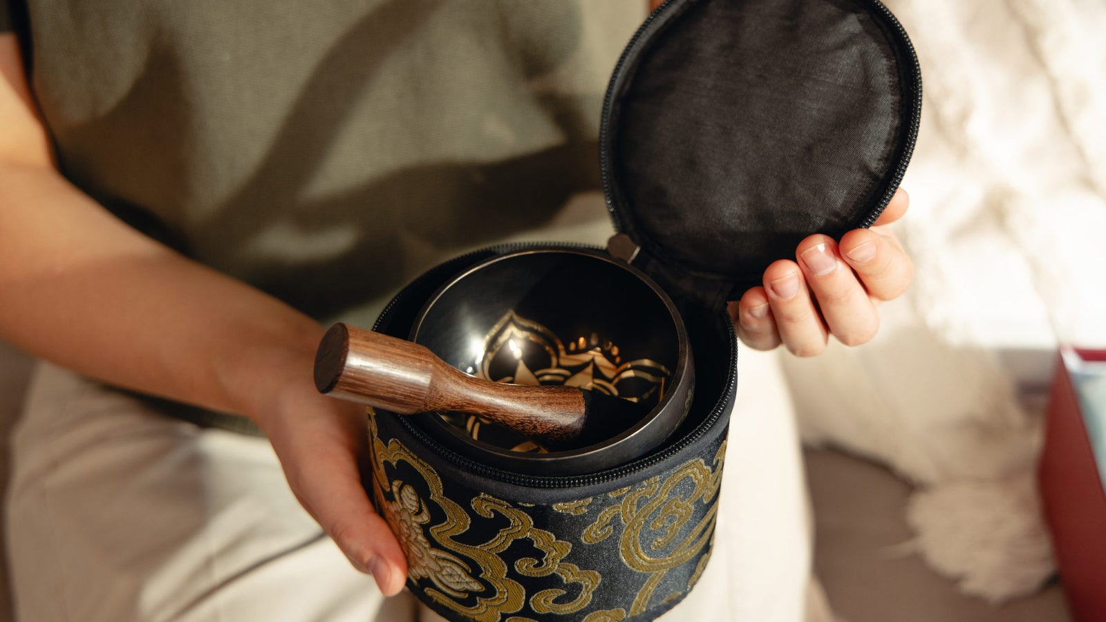 Hands lifting the lid of a brocade case with a singing bowl and striker packed inside
